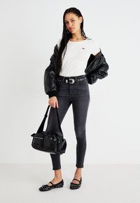 Mulher jovem com cabelo trançado, usando t-shirt branca, jaqueta de couro preta pendurada nos ombros, jeans cinzas, cinto com tachas e sapatos, segurando uma bolsa preta.