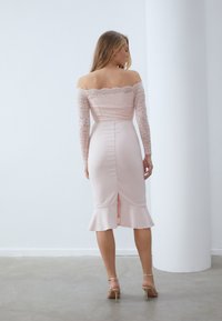 Robe rose décolletée avec des manches en dentelle, corsage ajusté et ourlet évasé. Présente une fente à l'arrière pour un détail supplémentaire.