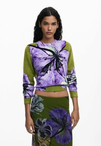 Trui met paars bloemenpatroon op groene stof, cropped ontwerp met lange mouwen. Gepaard met een groene rok met vergelijkbare bloemmotieven.