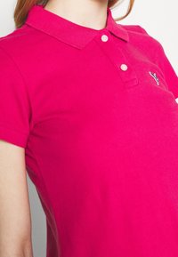 Rosa Poloshirt mit Kragen, kurzen Ärmeln und einer Knopfleiste; verfügt über ein kleines Logo im linken Brustbereich.