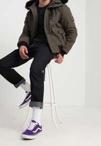 Olivengrøn oppustet jakke med sorte manchetter og pelsforet hætte, kombineret med mørke jeans og lilla Vans-sneakers. Hvide sokker er synlige.