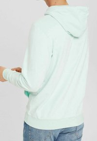 Sudadera con capucha en verde menta, textura suave, puños y dobladillo elásticos, bolsillo tipo canguro en la parte delantera, vista trasera con detalles de costura sutiles.