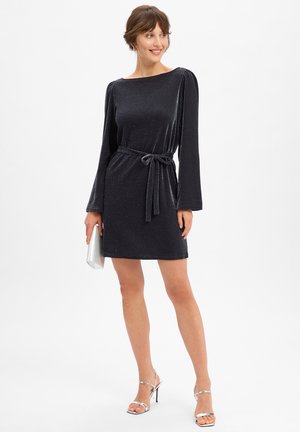 Femme portant une robe noire longue à manches longues ceinturée, tenant une pochette argentée, associée à des sandales à talons hauts argentées, debout sur fond blanc.