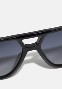 Ochelari de soare negri cu o ramă cu un design larg și un detaliu decupat la punte; lentilele gri reflectorizante reflectă lumina.