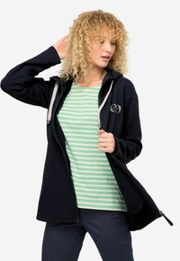 Giacca in pile blu navy con zip frontale, dettagli grigi e logo. Indossata sopra una maglietta a righe verde menta. Capelli ricci e look neutro.