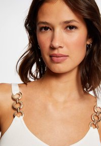 Top blanc à côtes avec des bretelles épaisses ornées d'accents en métal doré en forme de cercle. Texture lisse et décolleté en V. Maquillage naturel et cheveux de longueur moyenne.
