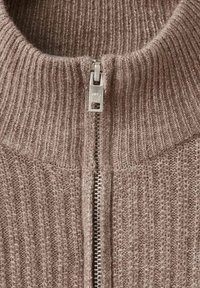 Primo piano di un maglione a costine marrone con una cerniera argentata parzialmente chiusa al colletto.