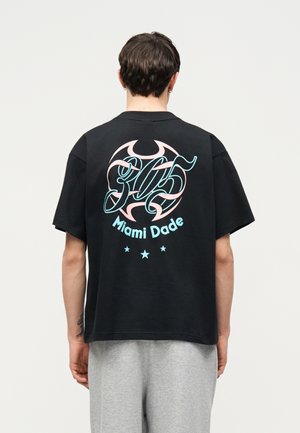 Person trägt ein schwarzes Oversized-T-Shirt mit pastellblauer und rosa "305 Miami Dade" Grafik und Sternen auf dem Rücken, kombiniert mit hellgrauen Hosen.