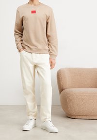 Felpa beige con logo rosso, pantaloni bianchi e sneakers bianche. Tessuto morbido e vestibilità casual; divano soffice sullo sfondo.