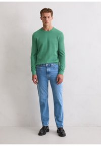 Grüner, gestrickter Langarmpullover, kombiniert mit hellblauen Jeans mit geradem Bein und schwarzen Lederschuhen. Schlichtes Design, mit gerippten Bündchen und Saum.