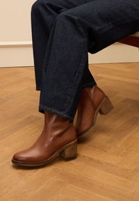 Braune Lederstiefeletten mit runder Spitze und gestapeltem Holzabsatz, kombiniert mit dunklen Jeans, auf einem Holzboden.