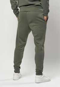 Persona che indossa pantaloni jogger verde oliva e sneaker bianche, in piedi con le mani nelle tasche, voltata di schiena contro uno sfondo semplice.
