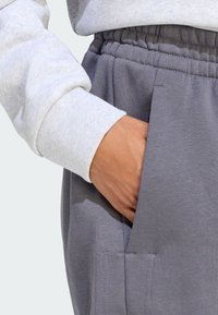 Pantaloni della tuta grigi con vita regolabile e tasche laterali, abbinati a un maglione grigio chiaro. Il tessuto appare morbido, testurizzato e informale.