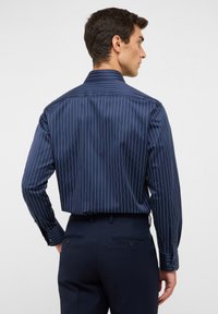 Camicia a righe navy realizzata in tessuto liscio, con colletto classico e maniche lunghe con polsini bottone, indossata con pantaloni scuri.