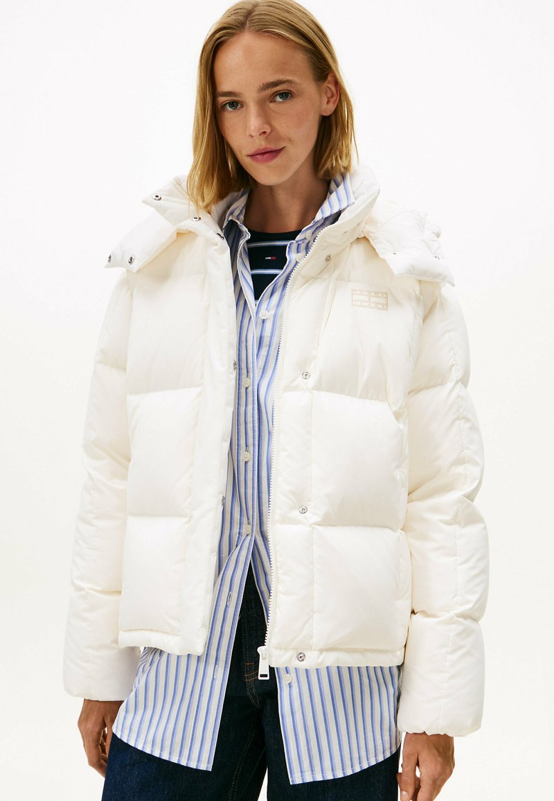 Chaqueta de plumas blanca con un diseño acolchado, cierre de cremallera y botones de presión, llevada sobre una camisa de rayas azul y blanca.