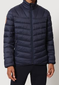 Uomo che indossa una giacca a vento trapuntata blu navy con colletto alto e tasche con zip, in piedi contro uno sfondo chiaro e uniforme.