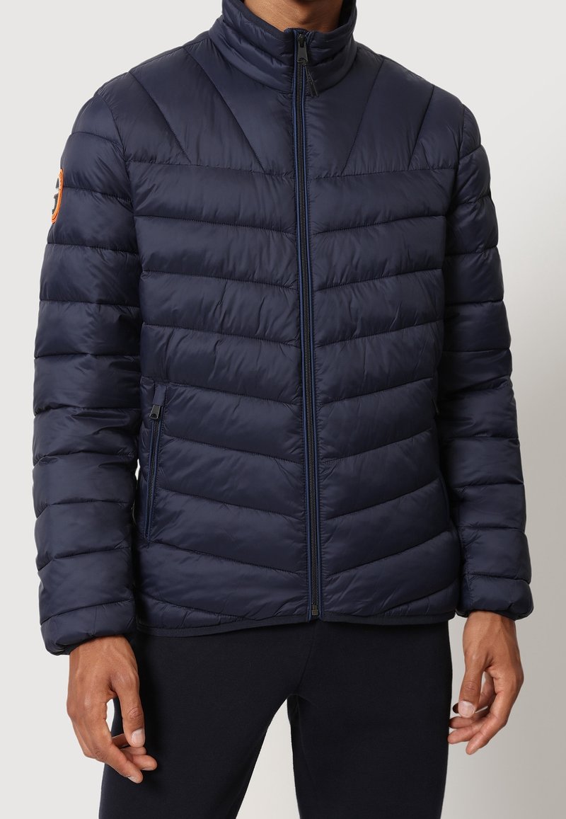 Uomo che indossa una giacca a vento trapuntata blu navy con colletto alto e tasche con zip, in piedi contro uno sfondo chiaro e uniforme.
