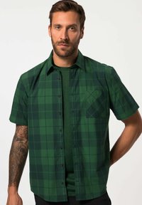 Camisa de cuadros verde de manga corta con botones, que presenta un bolsillo parchado y una camiseta de color verde oscuro debajo. Textura de tela suave.