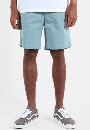 Homme portant un short bleu clair, un t-shirt blanc et des baskets grises avec des lacets blancs, debout les mains détendues le long du corps.