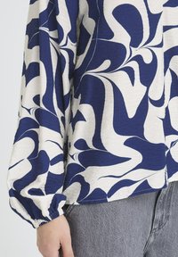 Locker sitzende Bluse aus strukturiertem beigem Stoff mit marineblauen abstrakten Wirbelmustern. Verfügt über elastische Bündchen und einen geschwungenen Saum.