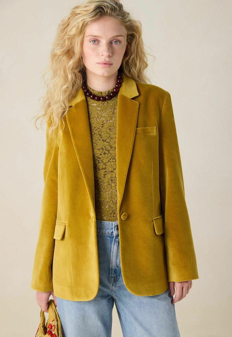 Blazer en velours jaune avec revers crantés, fermeture à un bouton et poches à rabat à l'avant, porté sur un haut en dentelle verte et un jean bleu.