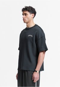 Jeune homme aux cheveux courts et bouclés porte un t-shirt noir texturé oversize et un short noir, debout de profil sur un fond blanc uni.