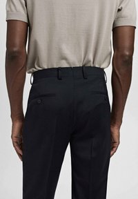 Pantalones negros con un corte ajustado, que cuentan con bolsillos laterales y una cintura sin costuras. La vista trasera muestra detalles de botones en los bolsillos.