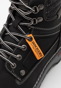 Bottes noires avec lacets gris, en cuir et en feutre. Comprend une étiquette logo orange et des œillets en métal. Accents de couture blanche présents.