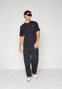 Musta lyhythihainen T-paita, jossa on yksinkertainen muotoilu ja pieni logo, yhdistettynä tummaan cargo-housuihin ja valkoisiin tennareihin. Mallilla on aurinkolasit.