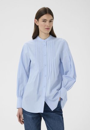 Vrouw draagt lichtblauwe geplooide blouse met knopen en lange opgeblazen mouwen, en donkerblauwe spijkerbroek, staand tegen een effen achtergrond.