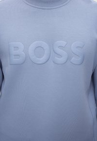 Ανοιχτό μπλε φούτερ με ανυψωμένα γράμματα τύπου μπλοκ που σχηματίζουν τη λέξη "BOSS" στο στήθος, ριμπ λαιμόκοψη και μακριά μανίκια.