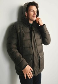 Ciemnozielona kurtka puchowa z kapturem, z zamkiem z przodu, dwiema przednimi kieszeniami i pikowanym wykończeniem. Model ma na sobie czarną koszulę pod spodem.