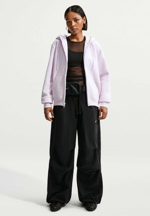 Junge Frau trägt ein helllila Zip-Up-Hoodie, transparentes schwarzes Oberteil, weite schwarze Hose, schwarze Bauchtasche und schwarze Sneaker und steht vor einem einfarbigen Hintergrund.