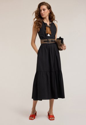 Un vestito nero senza maniche con una gonna a strati, accentuato da una cintura intrecciata marrone. Il modello tiene una pochette e indossa sandali con tacco quadrato rossi.