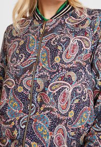 Marine bomberjakke med paisley mønster i pink, turkis og gul. Har guld lynlås og ribbet krave og manchetter. Struktureret stof.