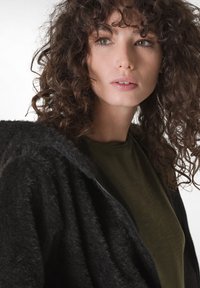 Giacca nera in pelliccia sintetica con zip e superficie testurizzata, abbinata a una maglietta verde scuro a maniche corte. Capelli ricci del modello parzialmente visibili.
