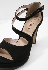 Sandale à talon haut en daim noir avec bout ouvert, brides croisées à la cheville et fermeture à boucle, semelle intérieure beige rembourrée avec logo de la marque.