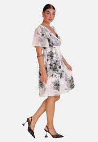 Vestido floral con escote en V, mangas cortas transparentes y cintura fruncida. Presenta un fondo blanco con patrones florales en gris y dorado. Zapatos de tacón negros.