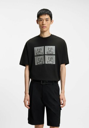 Jeune homme portant un t-shirt noir avec quatre inscriptions « Road 93 HUGO » et un short noir sur un fond blanc uni.