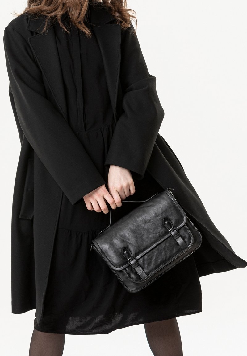 Femme portant un manteau et une robe noirs tenant un sac à main en cuir noir par sa poignée sur un fond blanc uni.