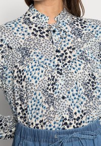 Blusa floreale di un delicato color crema con fiori blu e dettagli scuri. Presenta un colletto alto, chiusura con bottoni e una texture a pieghe.