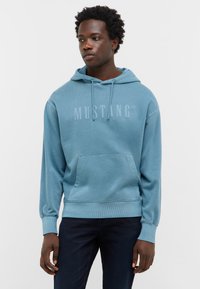 Haut à capuche bleu en coton, doté d'une poche avant et d'un logo "MUSTANG" en relief. Le modèle le porte avec un jean noir sur un fond uni.
