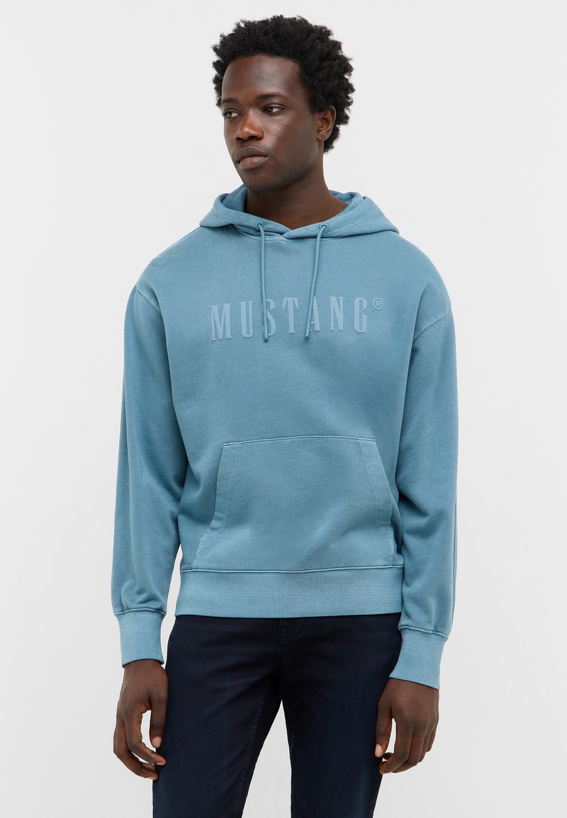 Haut à capuche bleu en coton, doté d'une poche avant et d'un logo "MUSTANG" en relief. Le modèle le porte avec un jean noir sur un fond uni.