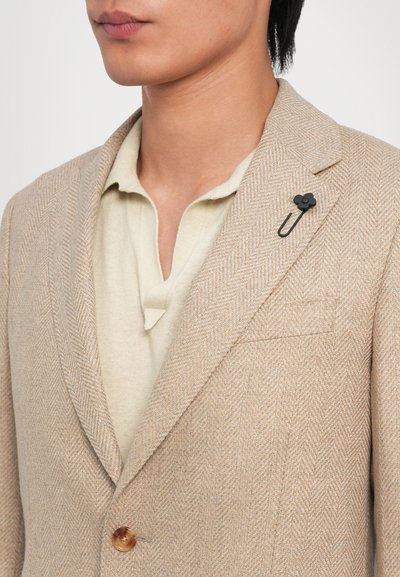 Beige Fischgrätenblazer mit Reverskragen, einknöpfiger Verschluss und einer schwarzen blumenförmigen Brosche am Revers. Hellgefärbtes Hemd darunter.