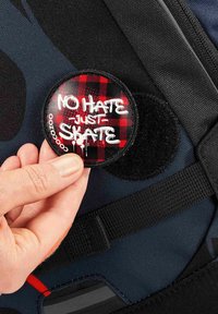 Runder Aufnäher mit rotem Karomuster, weißem Graffiti-Text „KEIN HASS, NUR SKATEN“, angebracht auf einer strukturierten dunklen Rucksackoberfläche.