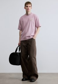 Ružové svetloružové tričko s krátkym rukávom a okrúhlym výstrihom s vyšívaným emblémom na hrudi, doplnené voľnými hnedými nohavicami a čiernou taškou.