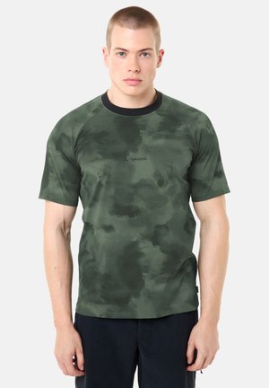 Uomo con capelli corti che indossa una maglietta a maniche corte Vaude con motivo mimetico verde e pantaloni scuri, in piedi davanti a uno sfondo chiaro e uniforme.