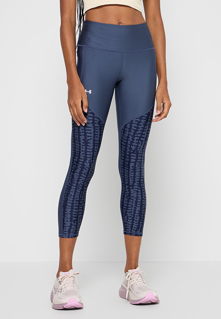 Mujer vistiendo leggings de Under Armour en azul marino con un patrón abstracto en la parte inferior de las piernas y zapatillas de correr rosa claro, de pie contra un fondo blanco.