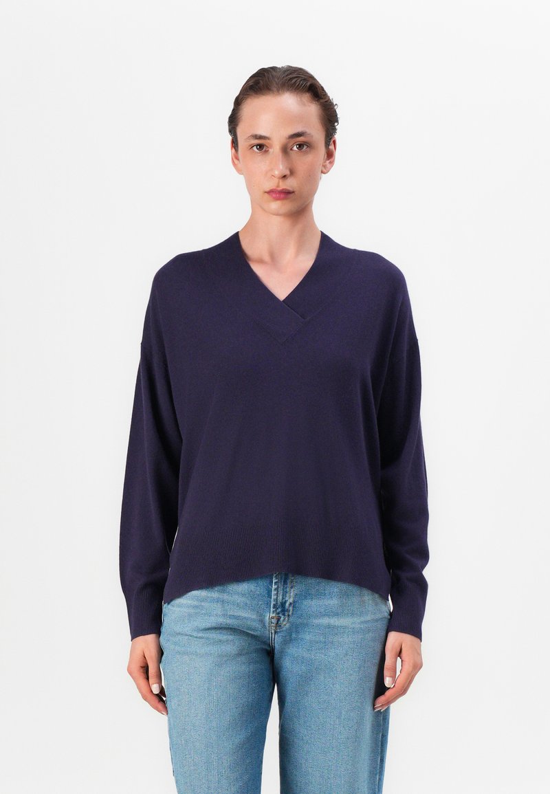 Pull à col en V, à manches longues, de couleur violet foncé, fabriqué à partir d'un tissu au toucher lisse. Il présente une coupe décontractée et un ourlet côtelé, associé à un jean bleu clair.