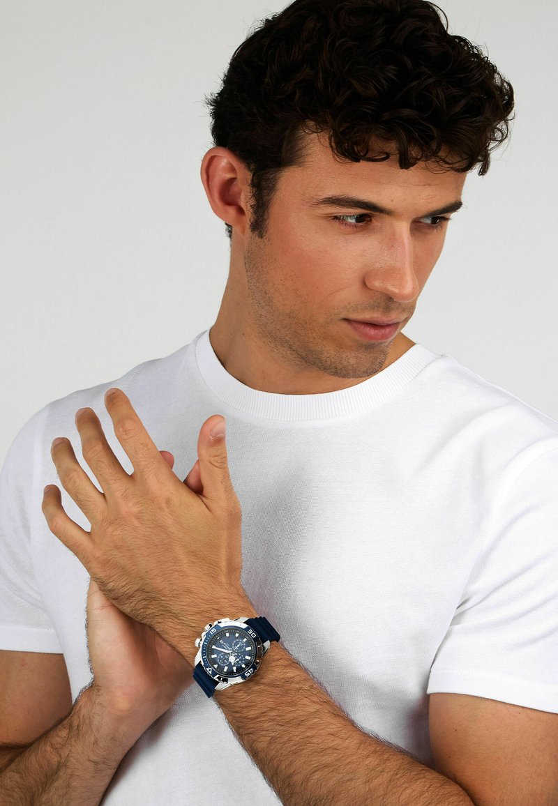 Orologio sportivo blu con case in acciaio inossidabile, quadrante blu scuro e cinturino in gomma. Modello che indossa una maglietta bianca semplice e mostra l'orologio.